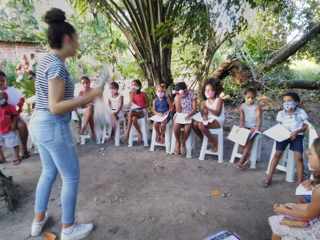 Outra imagem da Professora Thalia ministrando aula para os alunos que estão sentado e em círculo